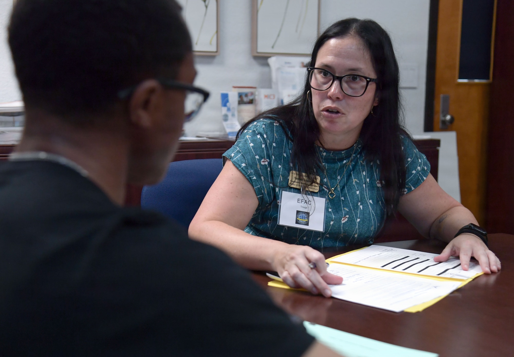 A woman speaks to a man from a table with a folder opened in front of her.