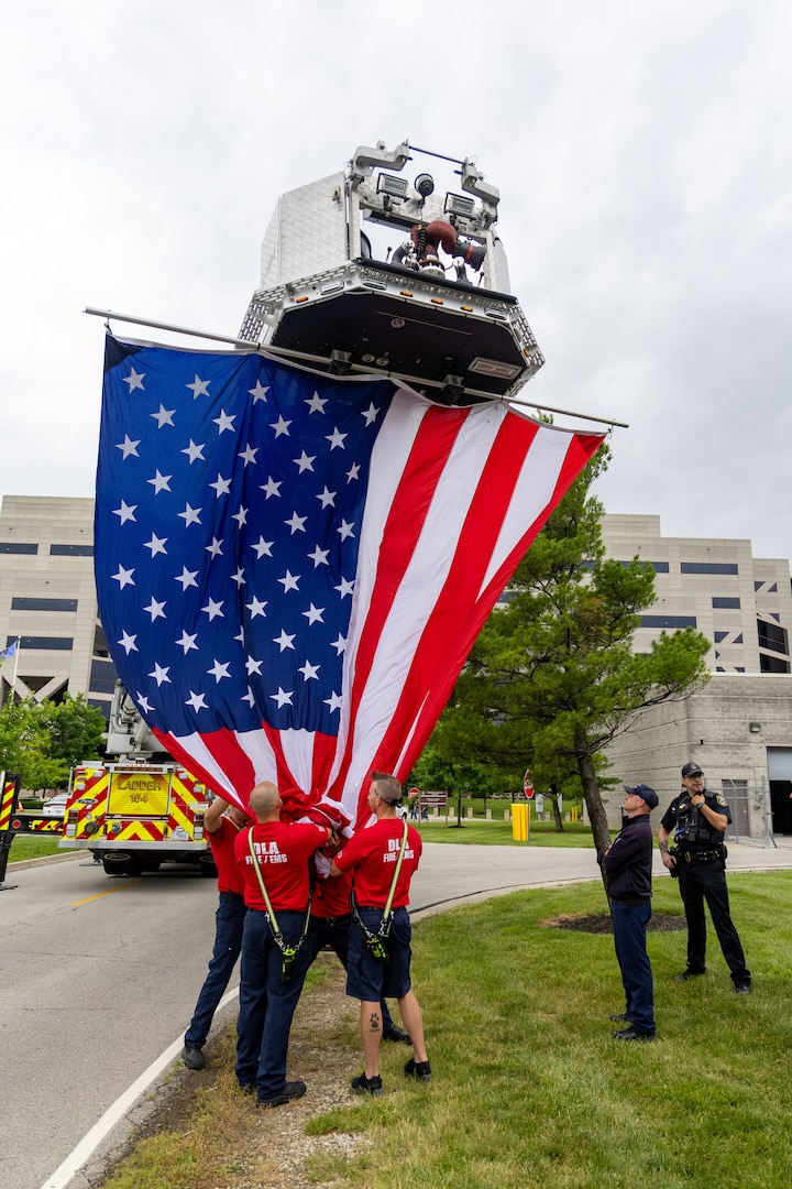 Several men in red shirts stand at base of flag as they raise it from fire truck ladder