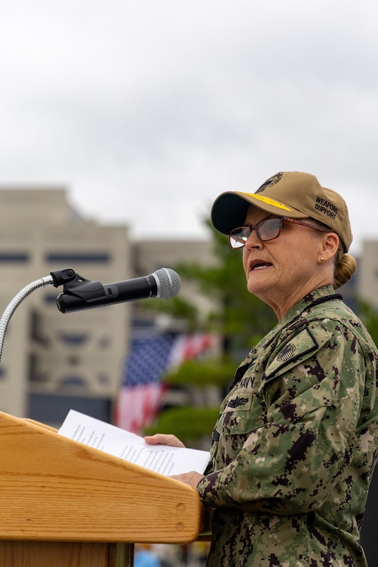 Woman in military uniform, glasses and tan ball cap stands at podium with microphone