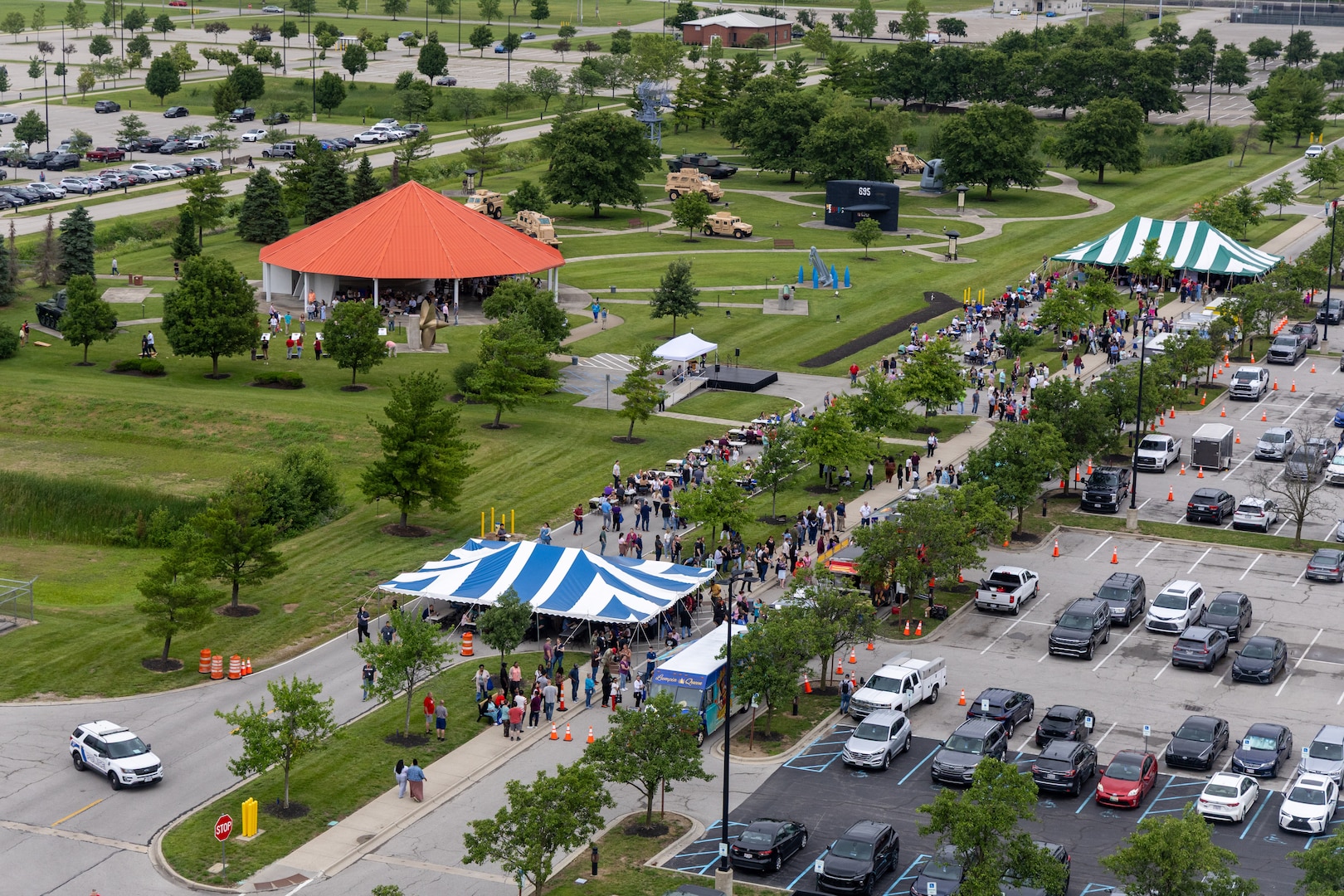 Overhead view of parking lot with two white and blue tents, lots of people and a pavilion with orange roof to the left