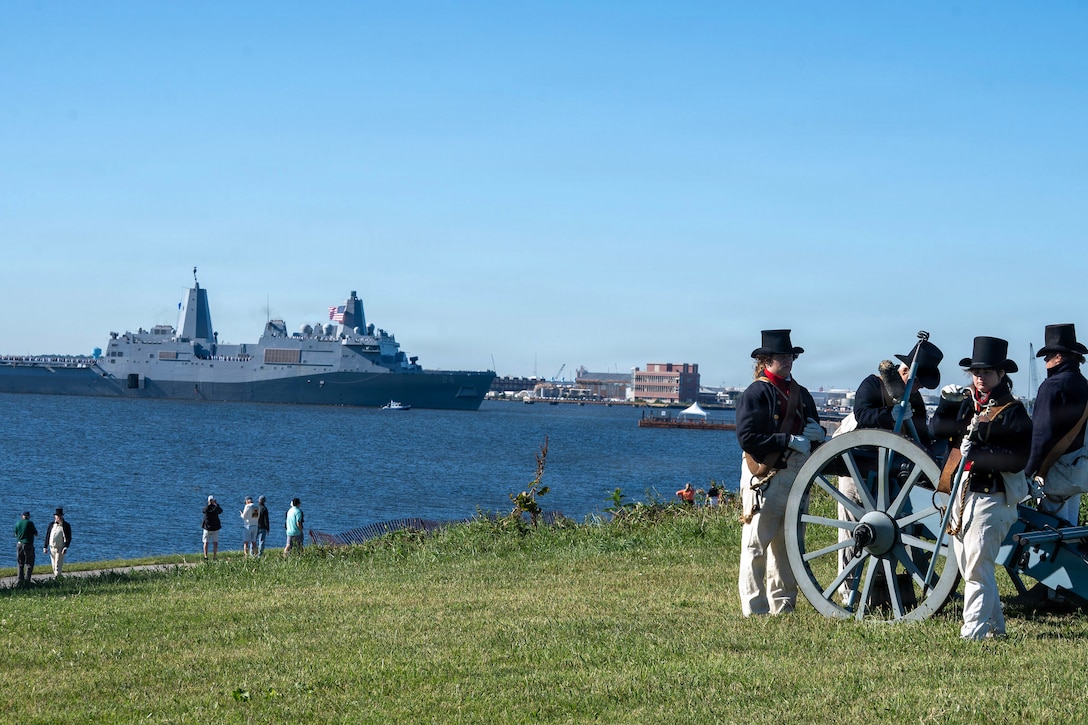 National Park Service personnel in ceremonial dress load a cannon in a field as spectators watch a naval ship transit a body of water during the day.