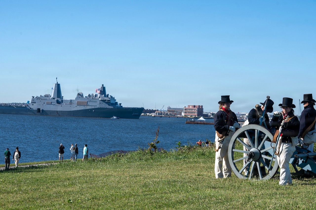 National Park Service personnel in ceremonial dress load a cannon in a field as spectators watch a naval ship transit a body of water during the day.