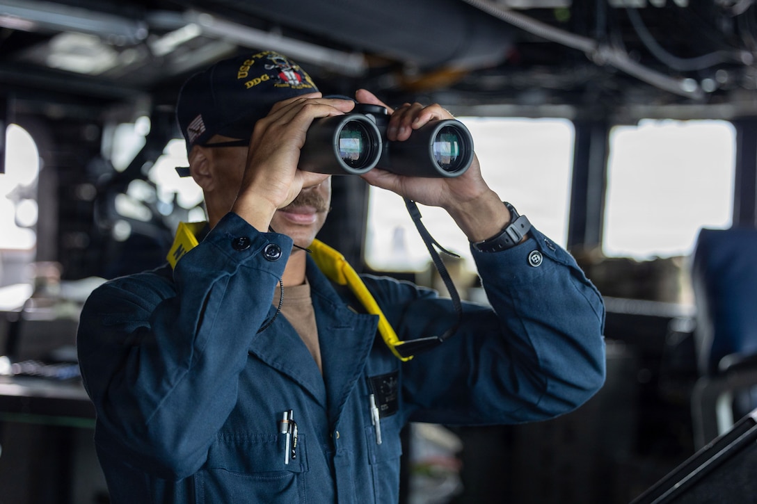 A sailor in a ship's pilot house uses binoculars to look out the window.