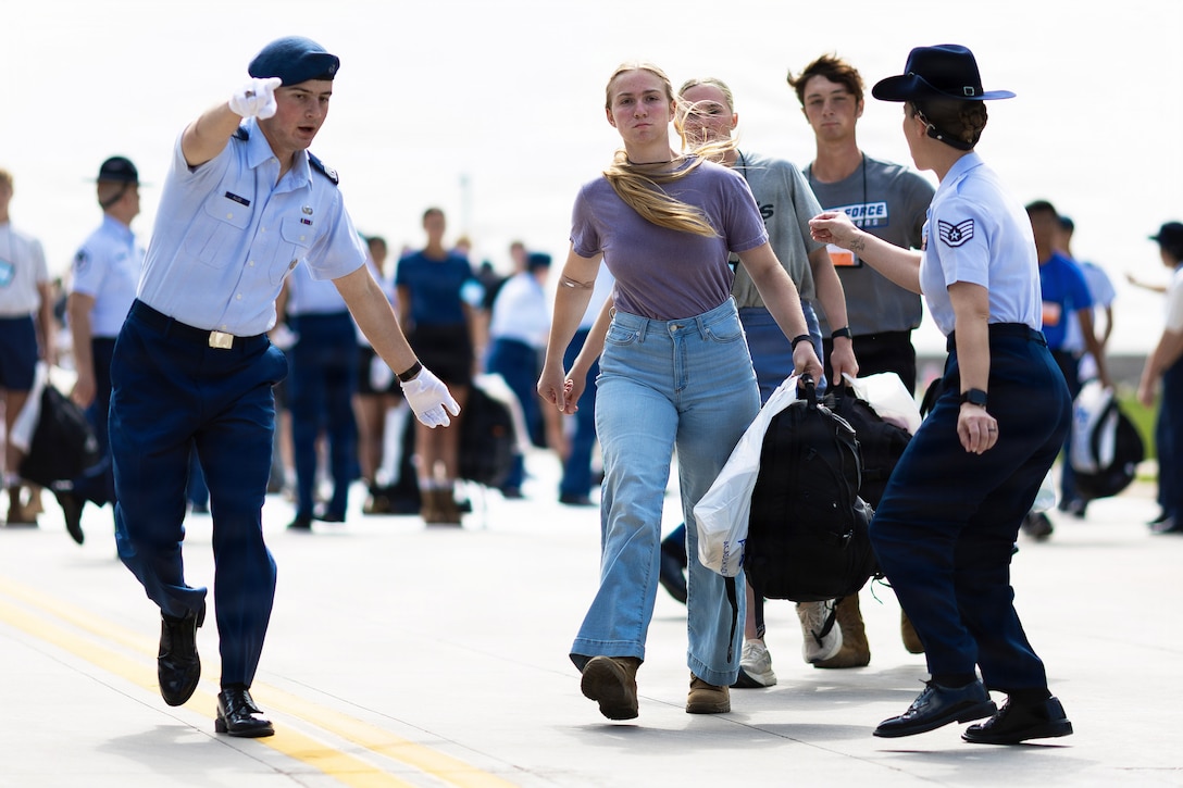 Three incoming members of the U.S. Air Force Academy walk through two service members giving them directions with a crowd in the background.