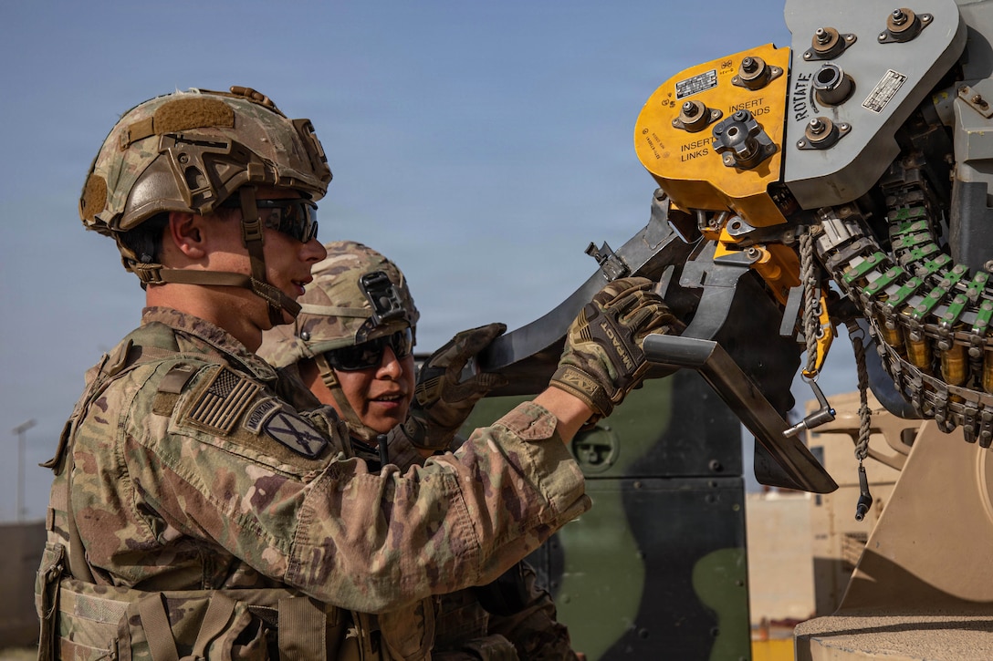 A soldier in tactical gear inspects a counter rocket, artillery and mortar system as another soldier watches against a blue sky.