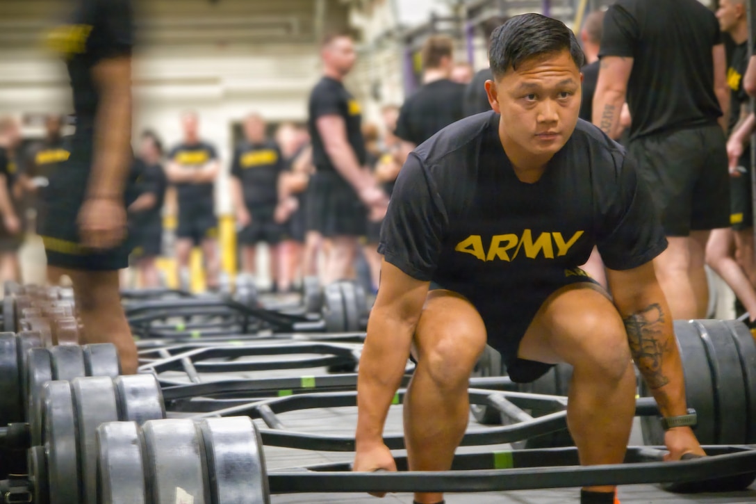A soldier in fitness attire squats to life heavy weights in a gym. Other soldiers and barbells with weights are in the background.