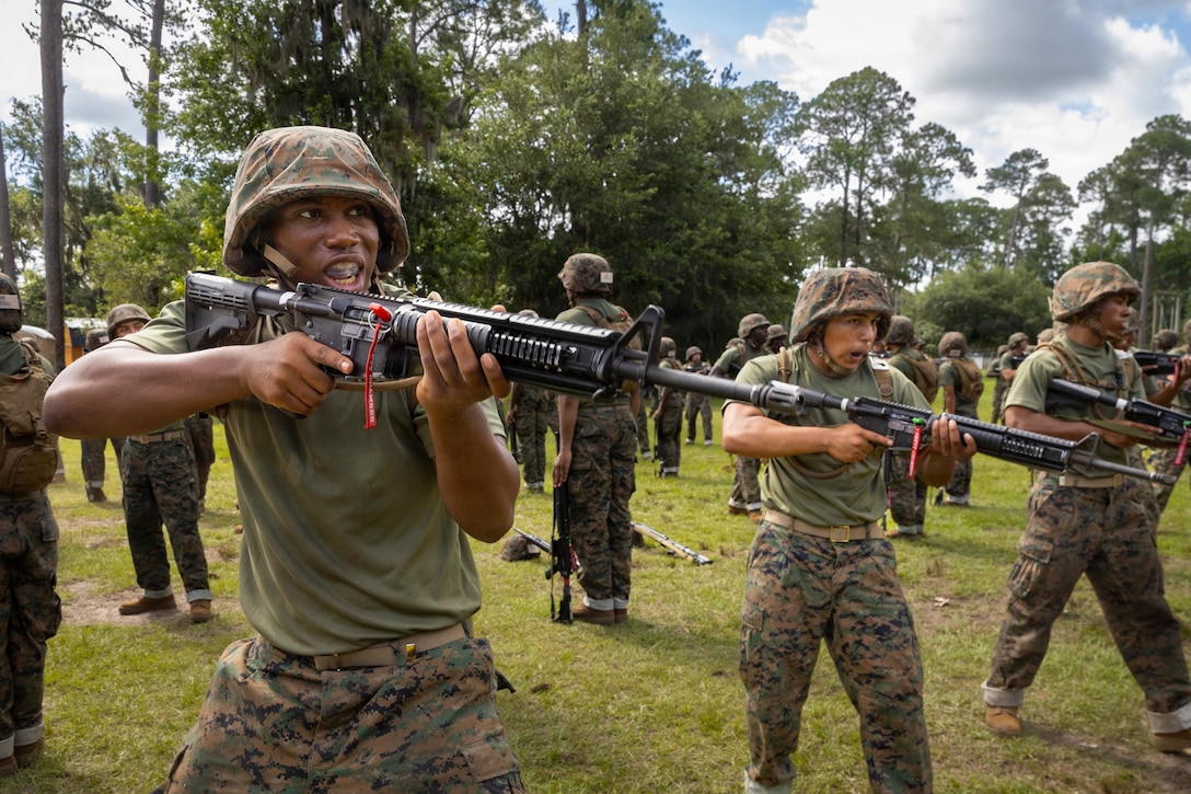 Marine recruits hold rifles outside and stand with legs apart and one in front of the other, as drill sergeants and other recruits stand in the background.