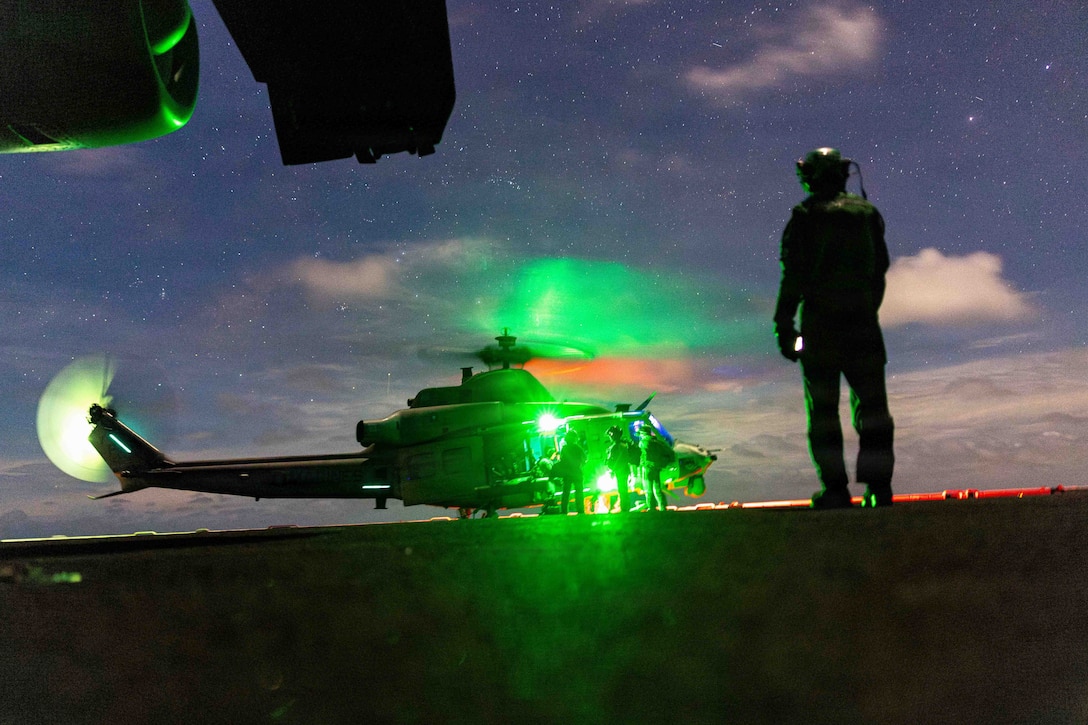 A Marine in silhouette looks at a helicopter on the ground a few hundred yards away under the stars. Green light is reflected on the helicopter.