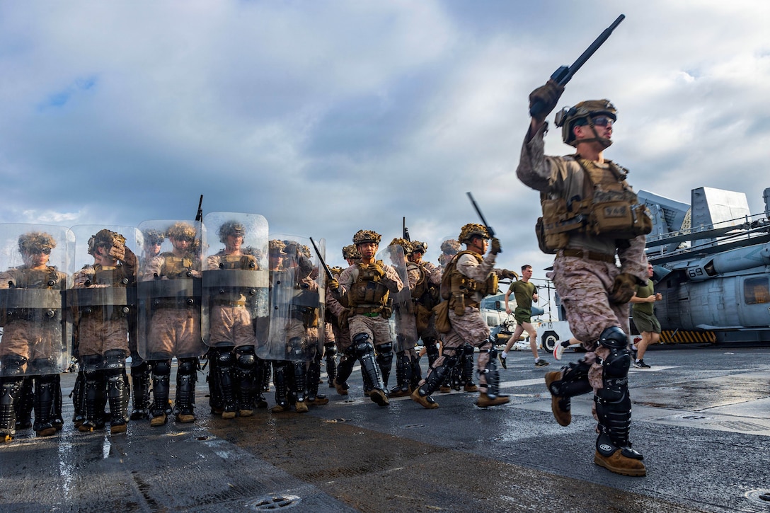 A group of Marines in camouflage and leg pads hold shields and weapons on the deck of a ship. One Marine stands in front of the group.