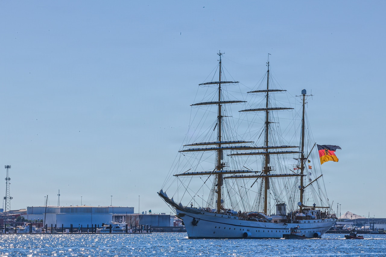 A large ship sails in a harbor with small tugboats guiding it; the ship is flying the German flag.