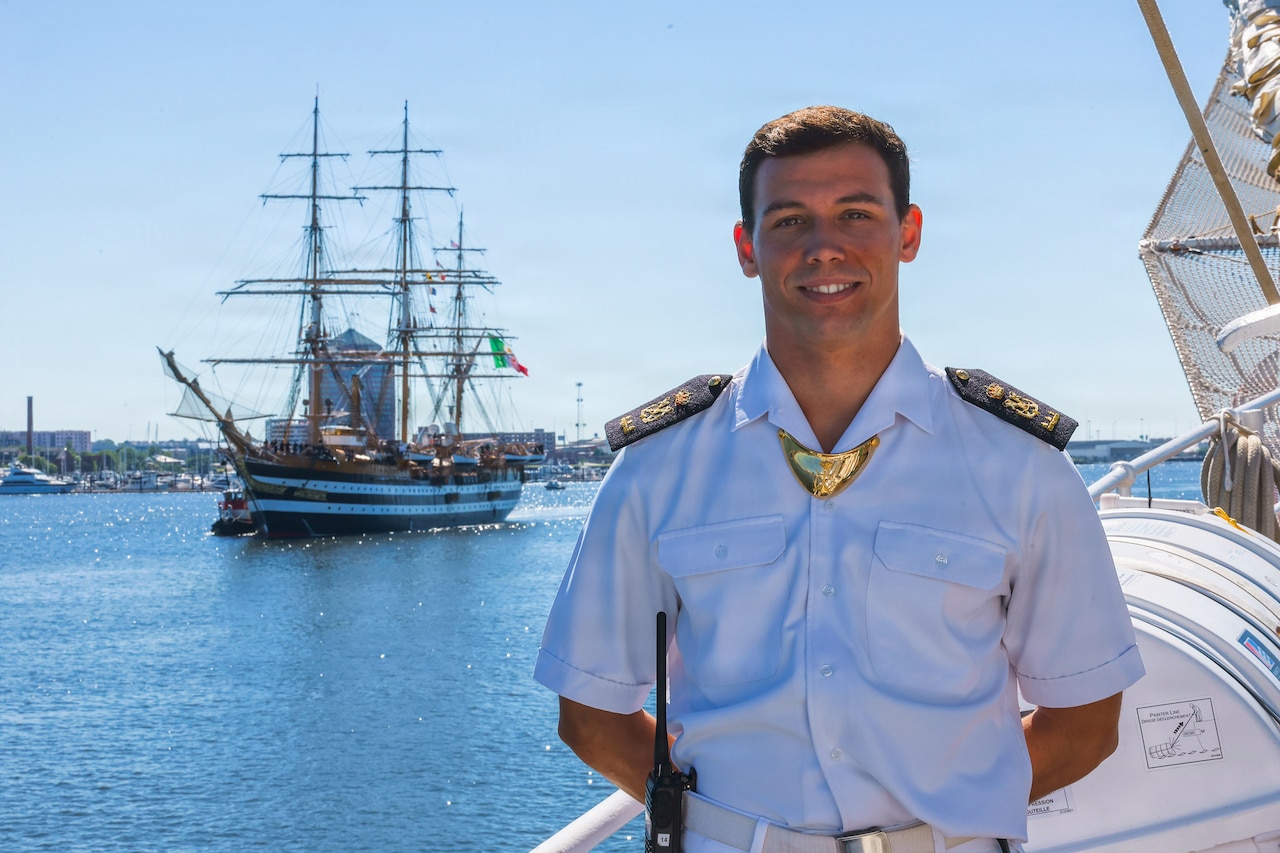 A man wearing a military dress uniform poses for a photo at a dock with a large ship sailing in a harbor in the background.