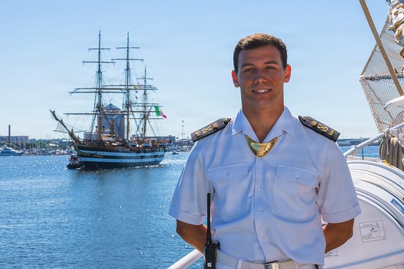 A man wearing a military dress uniform poses for a photo at a dock with a large ship sailing in a harbor in the background.