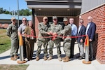 Photograph of a ribbon-cutting ceremony with military personnel and civilians standing in front of a brick building entrance. Three uniformed individuals are cutting a red ribbon stretched between two stanchions, symbolizing the opening or inauguration of a facility.