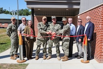 Photograph of a ribbon-cutting ceremony with military personnel and civilians standing in front of a brick building entrance. Three uniformed individuals are cutting a red ribbon stretched between two stanchions, symbolizing the opening or inauguration of a facility.