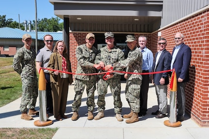 Photograph of a ribbon-cutting ceremony with military personnel and civilians standing in front of a brick building entrance. Three uniformed individuals are cutting a red ribbon stretched between two stanchions, symbolizing the opening or inauguration of a facility.
