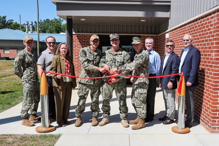 Photograph of a ribbon-cutting ceremony with military personnel and civilians standing in front of a brick building entrance. Three uniformed individuals are cutting a red ribbon stretched between two stanchions, symbolizing the opening or inauguration of a facility.