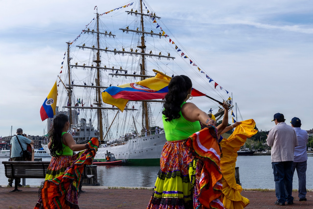 Four women wearing colorful dresses dance at a dock while a large ship sails in the nearby water; two of the women are waving Colombian flags.