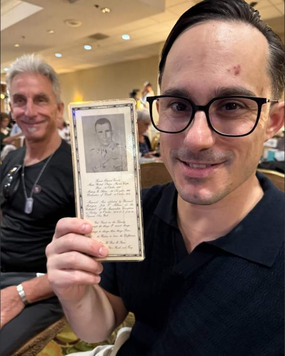 A man holds up a leaflet with a photo of a man and some cursive writing while another man looks on.
