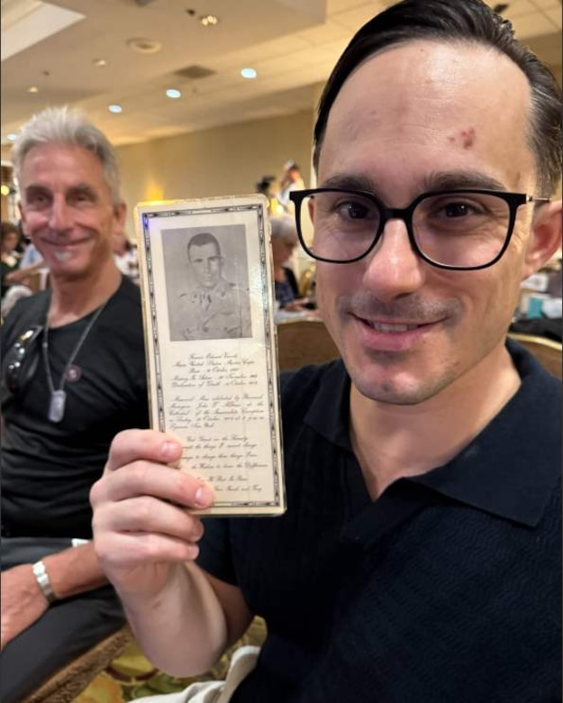 A man holds up a leaflet with a photo of a man and some cursive writing while another man looks on.