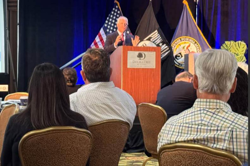 A man standing behind a lectern on a stage speaks to an audience.