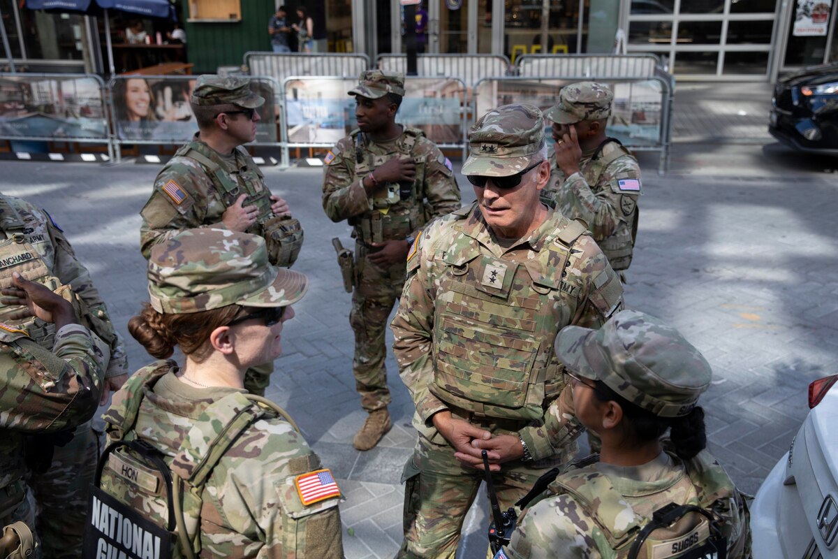 A man in a camouflage military uniform stands outside on a street speaking with two women in similar attire; five other people, also in camouflage military uniforms, are in the background.