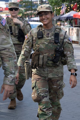 A woman in a camouflage military uniform and tactical gear smiles while walking down a city street. A man in similar attire walks behind her.