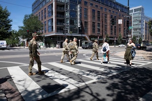 Five men in camouflage military uniforms and two women in casual attire cross a city street.