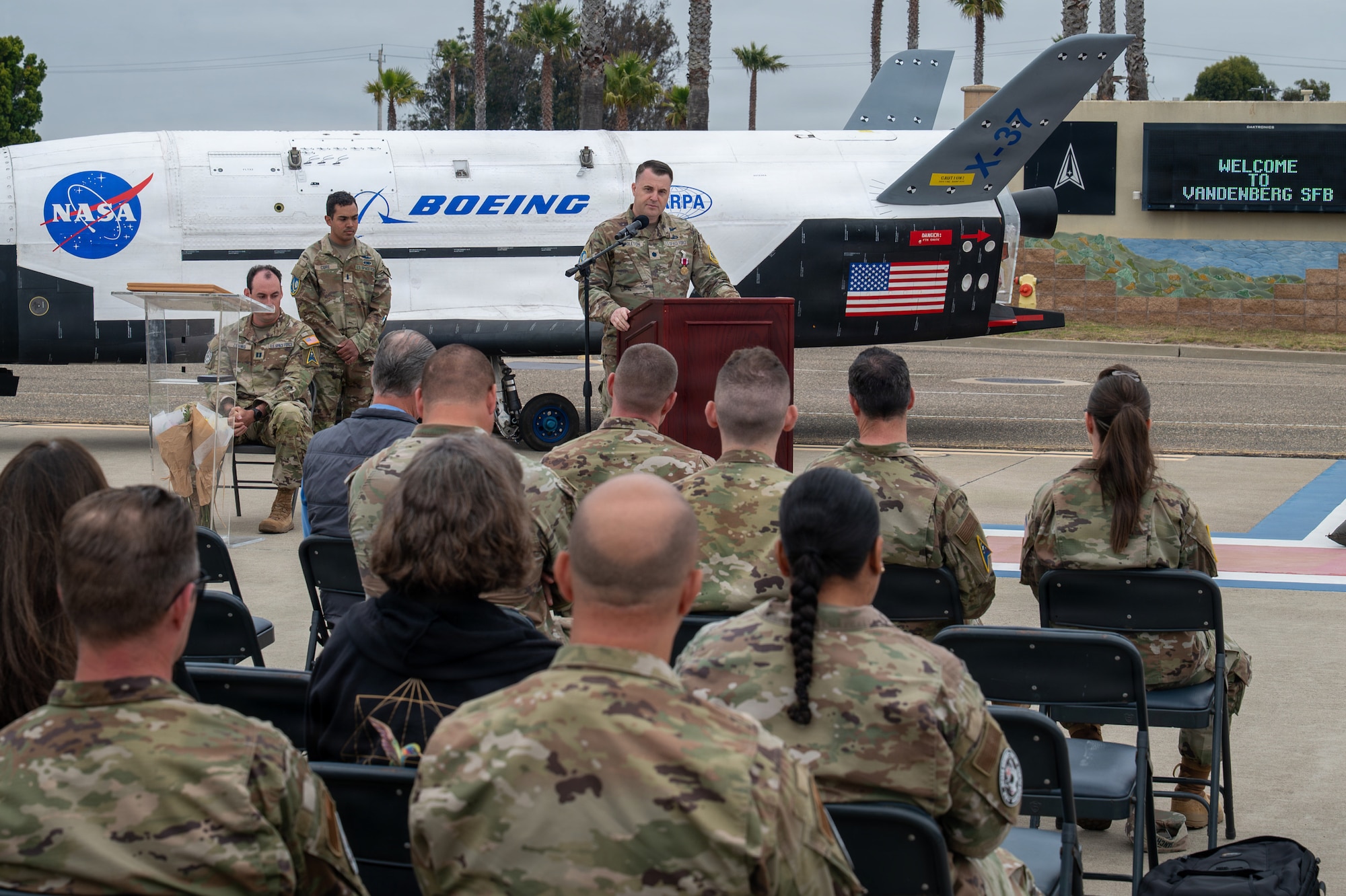 A person speaks from behind a podium while an audience sits and listens outside