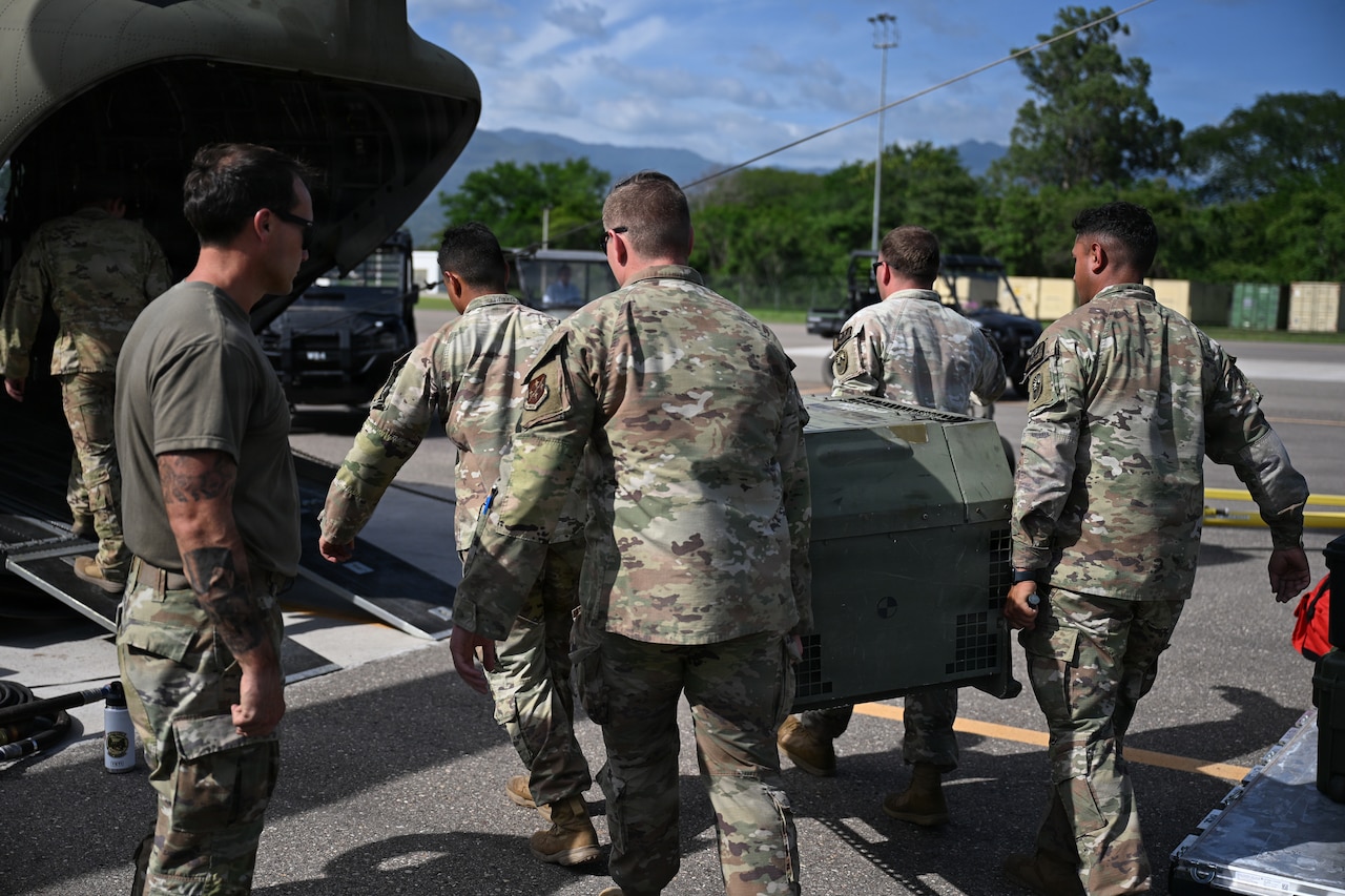 Four men wearing camouflage military uniforms carry a box into the back of a military cargo airplane. Another man in a camouflage military uniform stands by and watches.