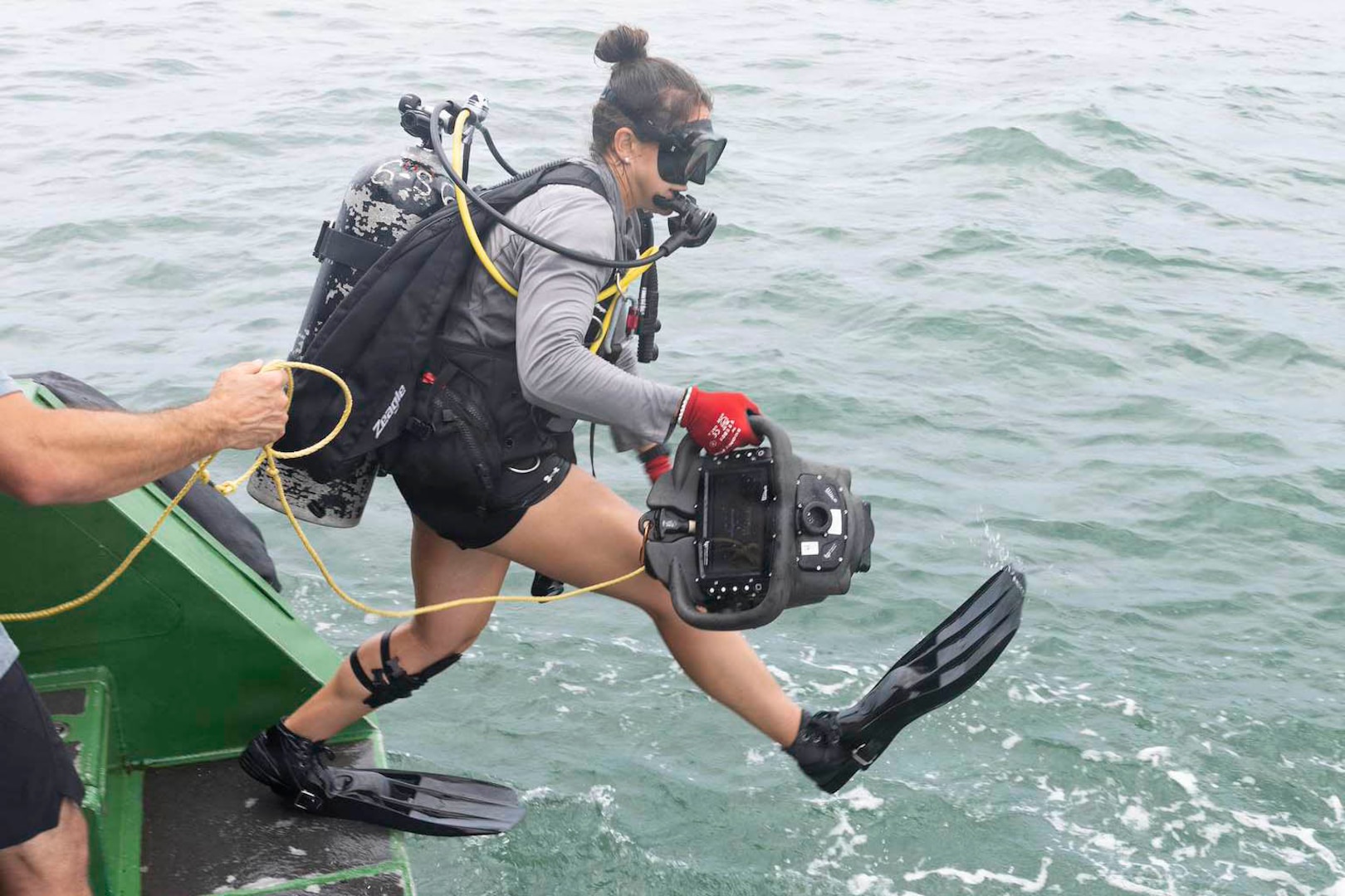 A woman wearing scuba gear jumps out of a boat into the ocean while holding equipment.