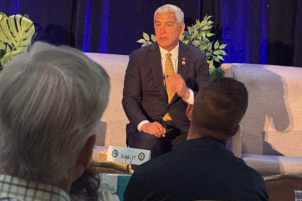A man in business attire sits in a chair on a stage and speaks to an audience.