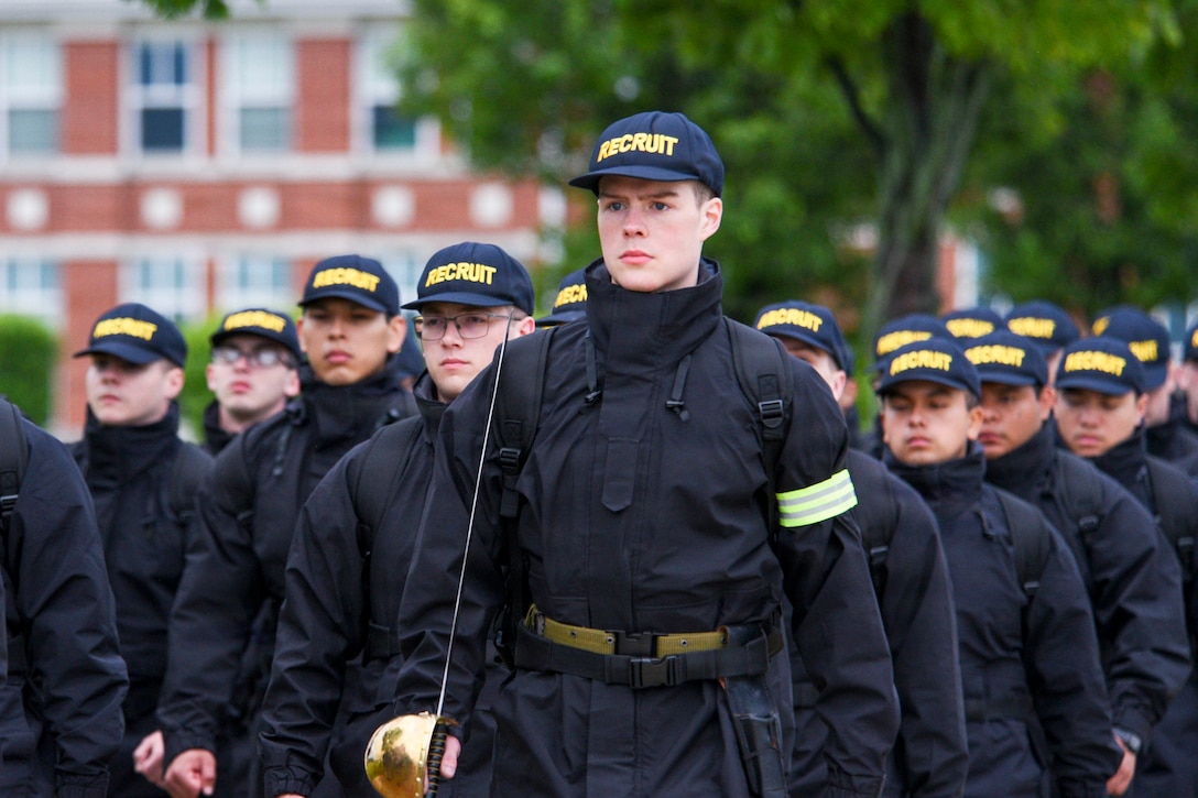 A Navy recruit carrying a sword leads fellow recruits wearing black uniforms and hats that say "recruits" as they march in formation in front of a blurred building and trees.