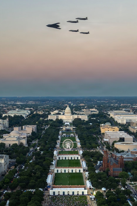 A U.S. Air Force B-2 Spirit leads four F-35A Lightning II aircraft during a formation flyover above the National Mall during a Freedom 250 event with pink yellow blue sky.