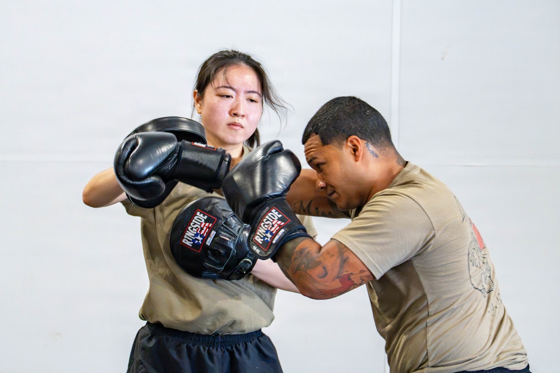 A soldier wearing boxing gloves hits a soldier wearing punching mitts against a white wall.