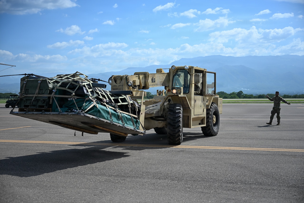 A man wearing a camouflage military uniform directs a forklift with supplies loaded onto a pallet. Mountains can be seen in the distance.