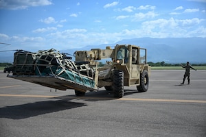 A man wearing a camouflage military uniform directs a forklift with supplies loaded onto a pallet. Mountains can be seen in the distance.