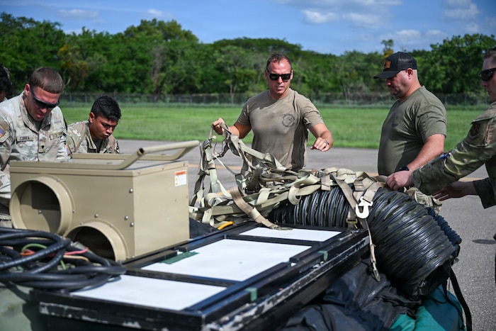 Five men wearing camouflage military uniforms load supplies onto a military cargo loader. A man in the middle of the five directs the others.