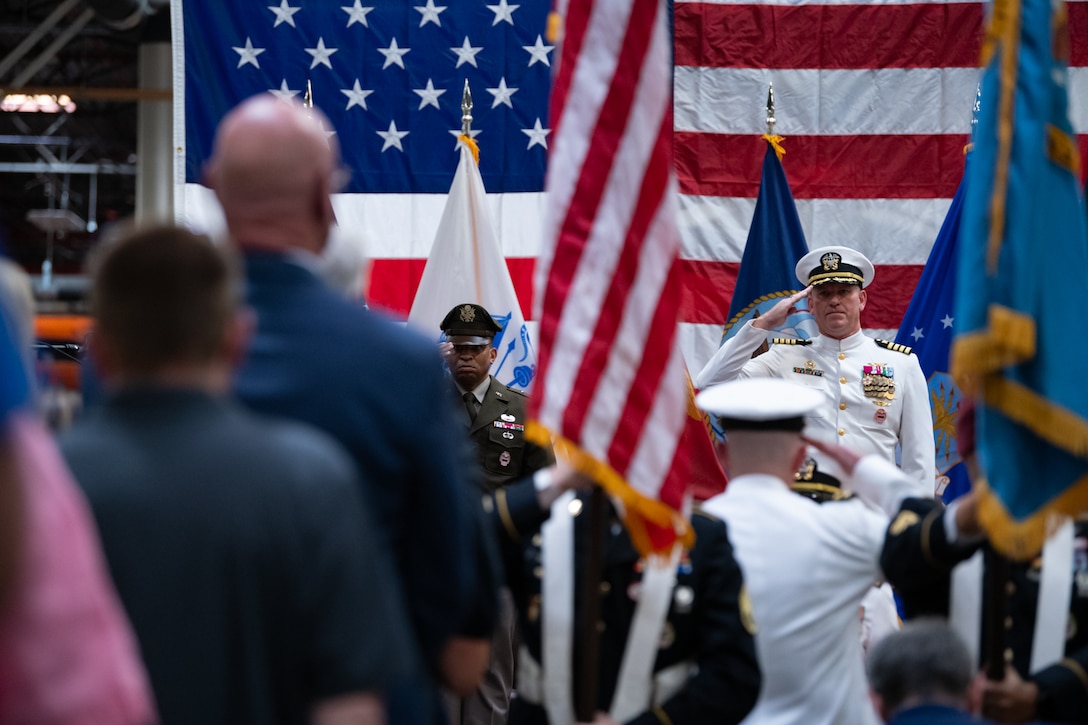 Two men wearing military uniforms stand on stage saluting flags