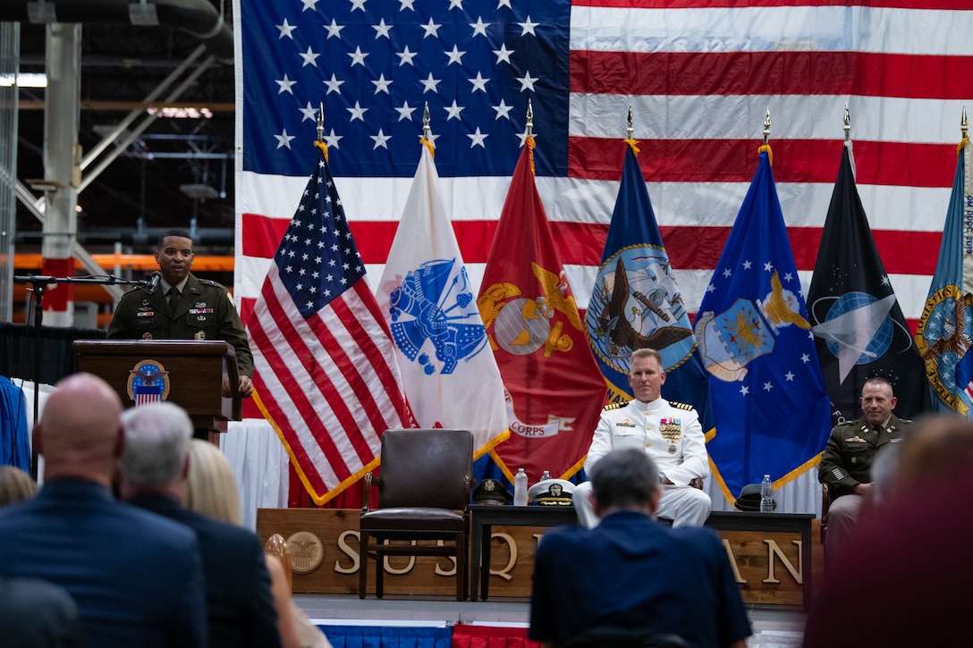 A man wearing a military uniform stands on stage behind a podium speaking into a microphone