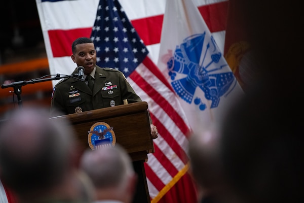 A man wearing a military uniform stands on stage behind a podium speaking into a microphone