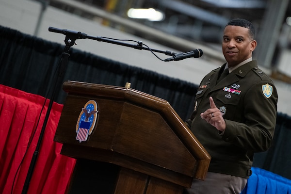A man wearing a military uniform stands on stage behind a podium speaking into a microphone