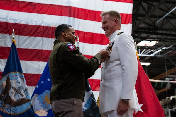Two men wearing military uniforms stand on stage, one is pinning a medal to the other's uniform.