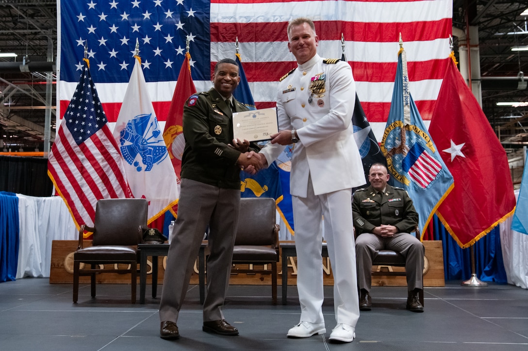 Two men wearing military uniforms stand on stage holding a certificate