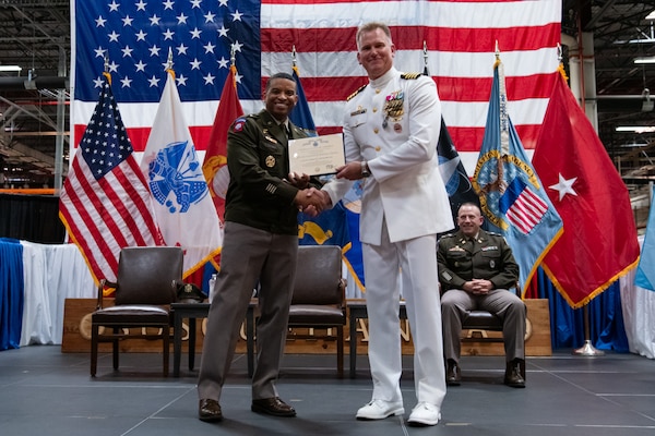 Two men wearing military uniforms stand on stage holding a certificate