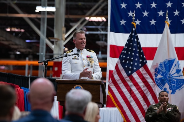 A man wearing a military uniform stands on stage behind a podium speaking into a microphone