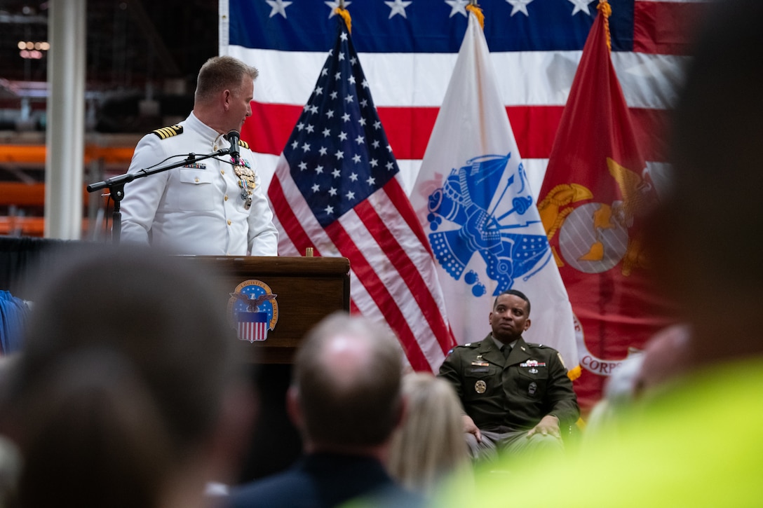 A man wearing a military uniform stands on stage behind a podium speaking into a microphone