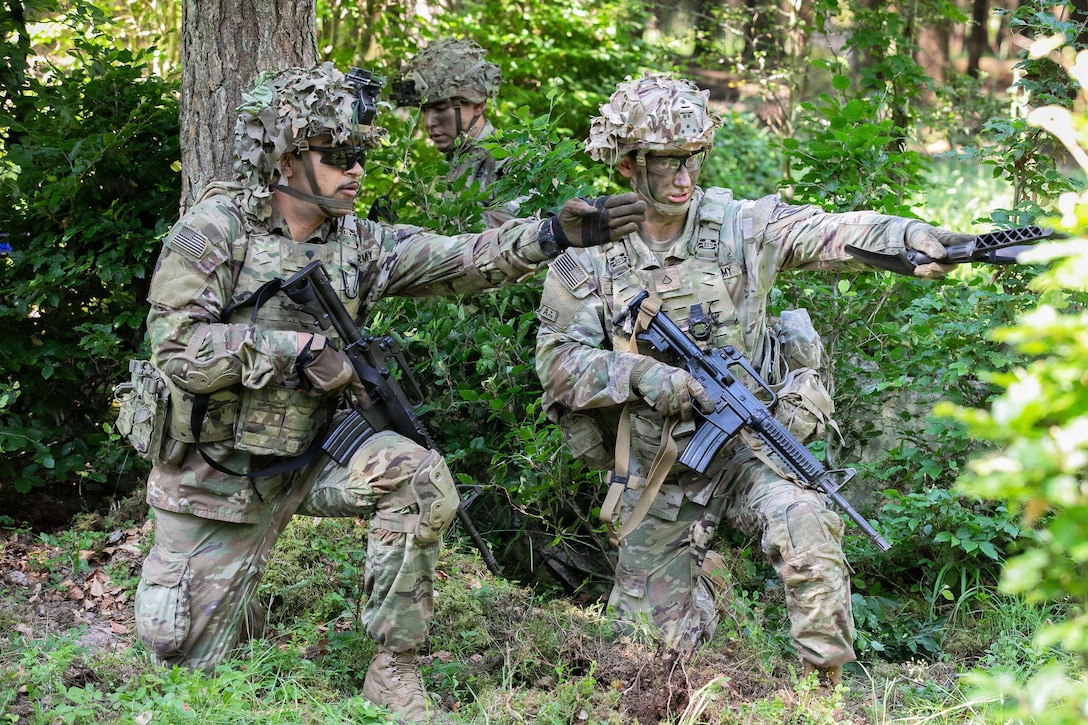 Two soldiers holding weapons each kneel on one knee and point an arm straight ahead. Another soldier is behind a bush in back of them.