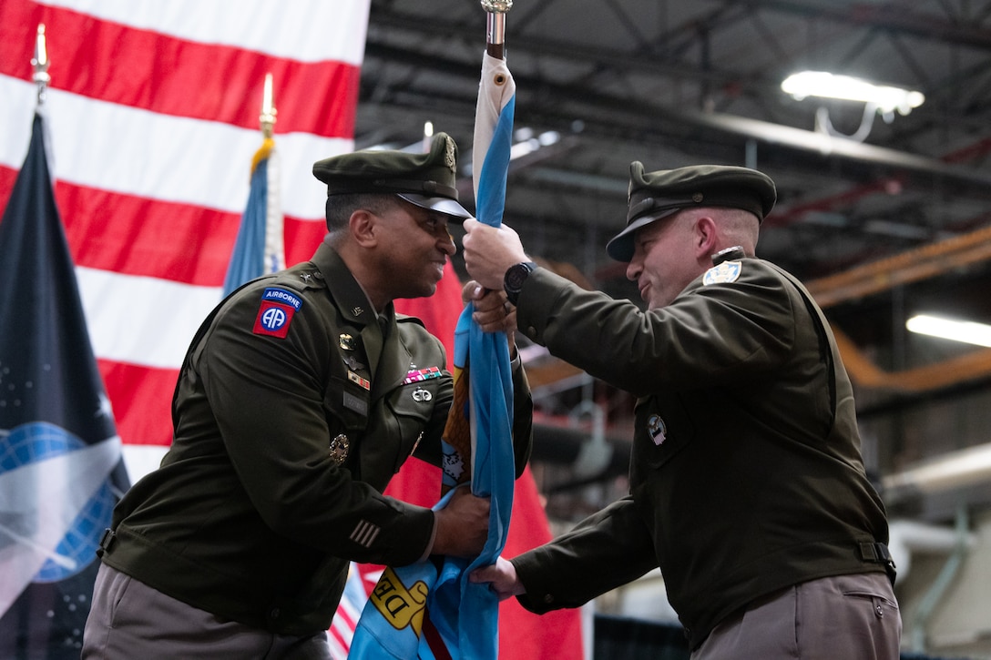 Two men wearing military uniforms pass a flag between one another.