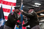 Two men wearing military uniforms pass a flag between one another.