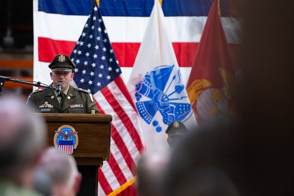A man wearing a military uniform stands on stage behind a podium speaking into a microphone